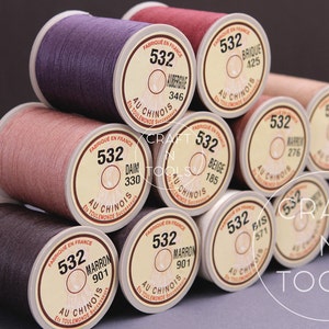May include: A close-up of a pile of spools of thread in various colors, including beige, brown, red, and purple. Each spool is labeled with the number "532" and the words "Au Chinois" in French. The spools are arranged in a pyramid shape.