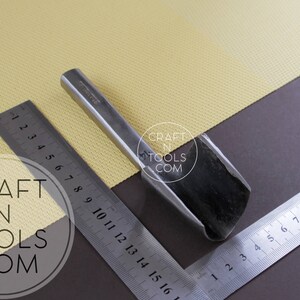 Puede incluir: Una herramienta de metal para trabajar el cuero con una cuchilla curva, que mide aproximadamente 15 cm de largo. La herramienta est&aacute; sobre una superficie marr&oacute;n con una regla.