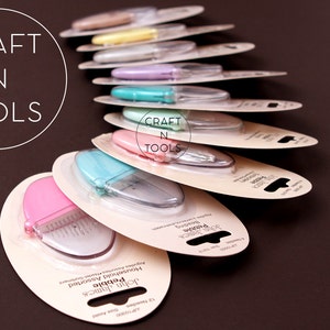 K&ouml;nnte beinhalten: Ein Stapel John James Pebble Haushalts-Scheren in verschiedenen Farben, darunter rosa, blau und gr&uuml;n, mit dem Markennamen und dem Produktnamen auf der Verpackung. Die Verpackung ist wei&szlig; mit schwarzem Text. Die Scheren sind in einer durchsichtigen Plastikverpackung verpackt.