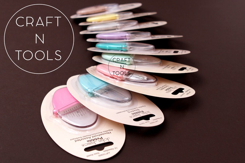 K&ouml;nnte beinhalten: Ein Stapel John James Pebble Haushalts-Scheren in verschiedenen Farben, darunter rosa, blau und gr&uuml;n, mit dem Markennamen und dem Produktnamen auf der Verpackung. Die Verpackung ist wei&szlig; mit schwarzem Text. Die Scheren sind in einer durchsichtigen Plastikverpackung verpackt.