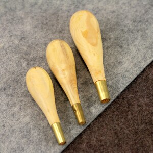 May include: Three wooden awls with brass tips, arranged in order of size from largest to smallest, on a gray and brown fabric background.