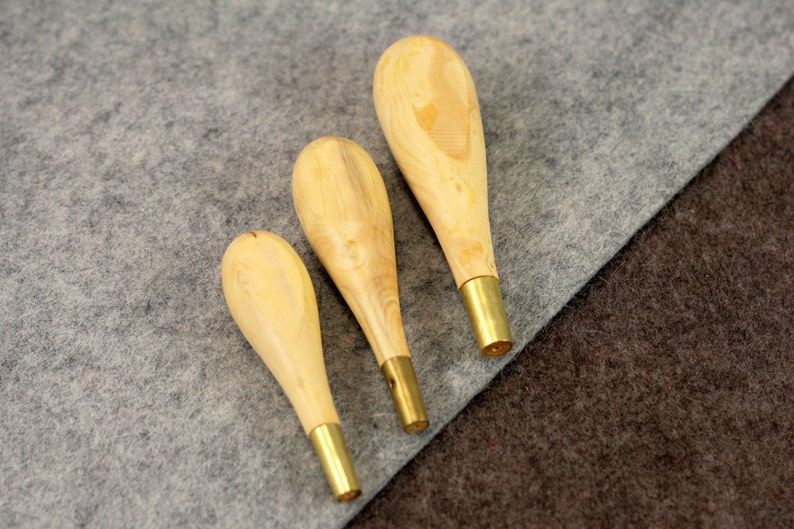 May include: Three wooden awls with brass tips, arranged in order of size from largest to smallest, on a gray and brown fabric background.