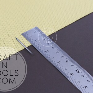 May include: A close-up of a silver needle and a ruler measuring 14 inches. The needle is lying on a yellow fabric with a grid pattern. The ruler has the text "CRAFT N TOOLS .COM" printed on it.