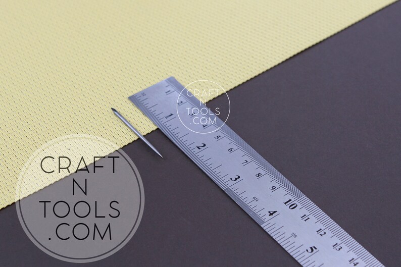 May include: A close-up of a silver needle and a ruler measuring 14 inches. The needle is lying on a yellow fabric with a grid pattern. The ruler has the text "CRAFT N TOOLS .COM" printed on it.