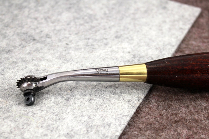 May include: A silver and brass leather stitching tool with a wooden handle. The tool has a star-shaped wheel and the text "VERGEZ" is engraved on the metal.