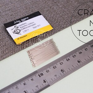 May include: A pack of 25 silver needles labeled "John James Saddiers Aiguilles Selkers Sattlermahnadeln L3911 Size 001" with a yellow background and a black and white illustration of a dragon. The pack is sitting on a gray fabric surface next to a silver ruler.