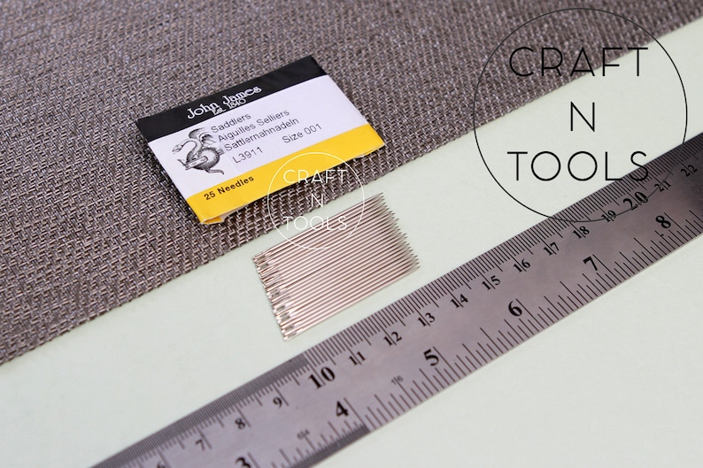 May include: A pack of 25 silver needles labeled "John James Saddiers Aiguilles Selkers Sattlermahnadeln L3911 Size 001" with a yellow background and a black and white illustration of a dragon. The pack is sitting on a gray fabric surface next to a silver ruler.