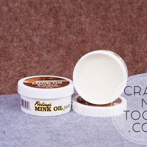 Pode incluir: Três potes brancos de pasta de óleo de vison Fiebing's, um condicionador para couro. Os potes estão empilhados em uma superfície marrom e cinza. O texto "CRAFT N TOOLS .COM" é visível no canto inferior direito da imagem.