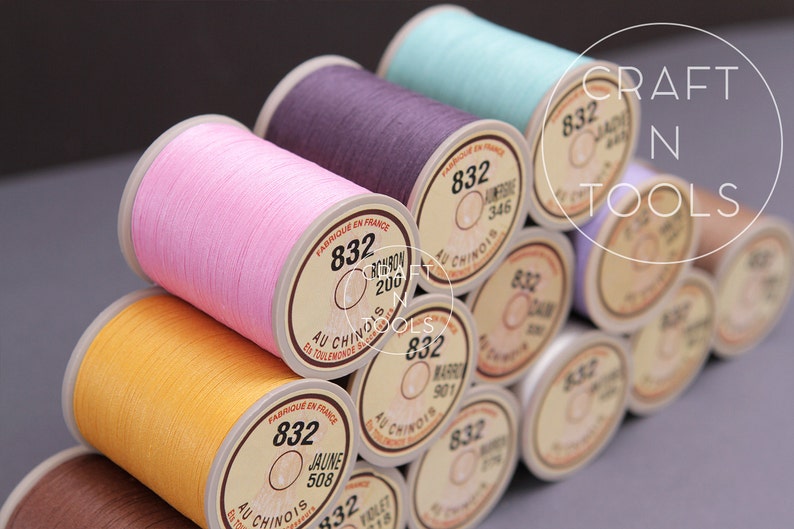 May include: A close-up of a stack of spools of thread in various colors, including pink, purple, yellow, and blue. The spools are labeled with the number "832" and the text "Au Chinois" in French. The spools are arranged in a pyramid shape.