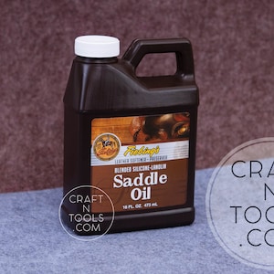 May include: A brown plastic bottle of Fiebings Saddle Oil with a white cap and handle. The label reads "Blended Silicone-Lanolin Saddle Oil" and "16 fl oz. 473 ml". The bottle is on a gray and brown background.