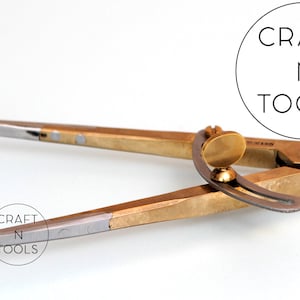 May include: A brass and silver compass with a curved handle and a pointed tip. The compass is designed for drawing circles and arcs. The compass is labeled "CRAFT N TOOLS".