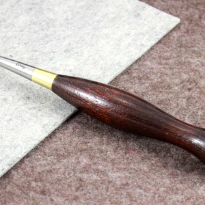 May include: A close-up of a leather stitching tool with a wooden handle. The tool has a curved metal blade with a small wheel at the end. The handle is dark brown and has a smooth finish.
