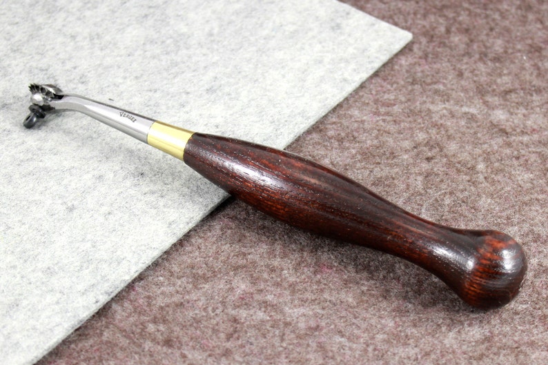 May include: A close-up of a leather stitching tool with a wooden handle. The tool has a curved metal blade with a small wheel at the end. The handle is dark brown and has a smooth finish.