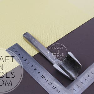 Puede incluir: Una herramienta de metal plateado con una cuchilla curva, utilizada para cortar cuero. La herramienta est&aacute; sobre una superficie marr&oacute;n con una regla y una tela amarilla.