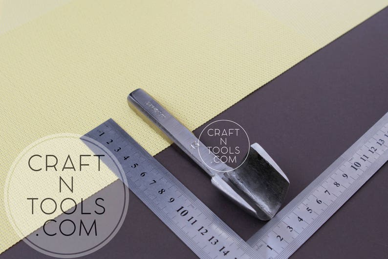 Puede incluir: Una herramienta de metal plateado con una cuchilla curva, utilizada para cortar cuero. La herramienta est&aacute; sobre una superficie marr&oacute;n con una regla y una tela amarilla.