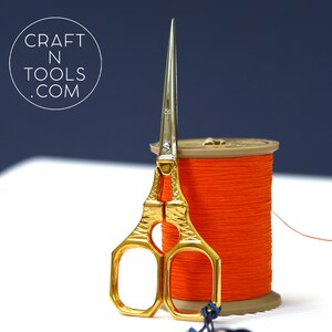 May include: Gold-coloured embroidery scissors with a sharp point and decorative handles, beside a spool of bright orange thread. The scissors are in front of the thread, with the text "CRAFT N TOOLS .COM" in a circle in the background.