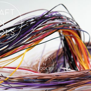 May include: A collection of colorful leather cords in various shades of purple, red, orange, yellow, brown, white, and black. The cords are arranged in a loose bundle, creating a textured and vibrant display.