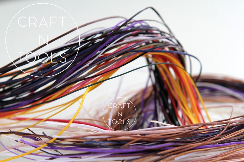May include: A collection of colorful leather cords in various shades of purple, red, orange, yellow, brown, white, and black. The cords are arranged in a loose bundle, creating a textured and vibrant display.