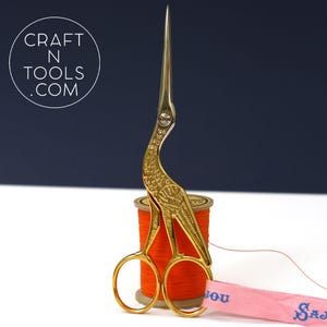May include: Gold-coloured scissors with a bird design, resting on a spool of orange thread. The scissors have a long, pointed blade and a curved handle. The spool of thread is sitting on a white surface.