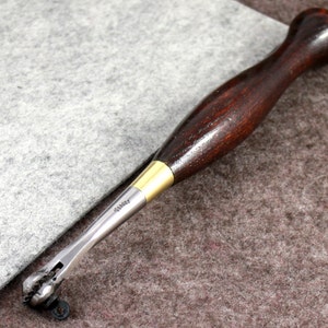 May include: A close-up of a leather stitching tool with a wooden handle and a metal stitching head. The tool is lying on a brown and gray surface.