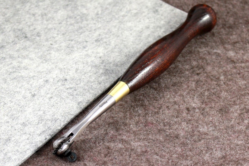May include: A close-up of a leather stitching tool with a wooden handle and a metal stitching head. The tool is lying on a brown and gray surface.