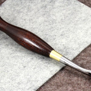May include: A wooden-handled leather sewing awl with a brass ferrule and a rotating wheel at the tip. The awl is lying on a gray fabric surface.