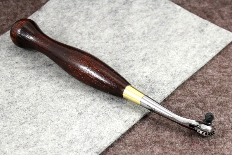 May include: A wooden-handled leather sewing awl with a brass ferrule and a rotating wheel at the tip. The awl is lying on a gray fabric surface.