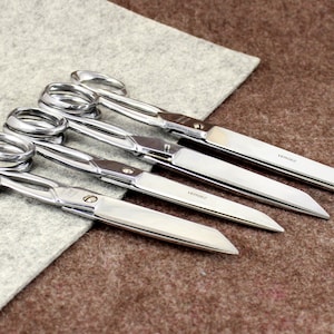 Puede incluir: Cuatro tijeras plateadas con cuchillas largas y mangos curvos. Las cuchillas son afiladas y los mangos son cómodos de sujetar. Las tijeras están dispuestas en una fila, con las tijeras más grandes en la parte superior y las tijeras más pequeñas en la parte inferior.