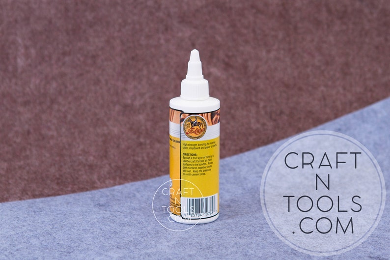 May include: A clear bottle of Leathercraft  Leather Cement with a yellow label. The label has a picture of a horse and the text "Leathercraft Cement" and "High strength bonding for leather, vinyl, shellac and paper crafts".