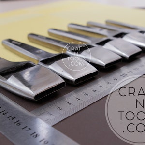 May include: A set of polished metal leather working tools with rectangular cutting heads, arranged on a brown surface with a metal ruler. The tools are angled towards the viewer. The text "CRAFT N TOOLS .COM" is visible.