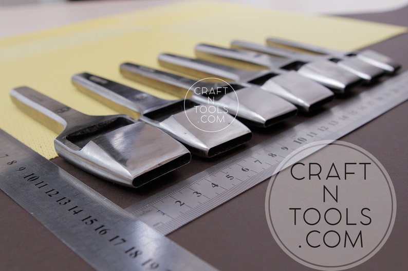 May include: A set of polished metal leather working tools with rectangular cutting heads, arranged on a brown surface with a metal ruler. The tools are angled towards the viewer. The text "CRAFT N TOOLS .COM" is visible.