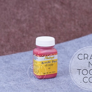 May include: A small bottle of Fiebing's Acrylic Dye for Leather in red. The bottle is labeled with the brand name, product name, and color. The bottle is sitting on a gray surface.