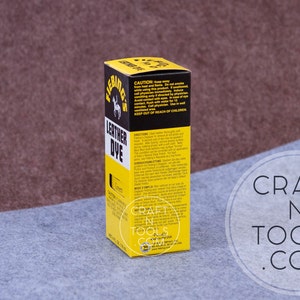 May include: A yellow and black bottle of Fiebing's Leather Dye. The bottle is labeled with the product name, color, and instructions for use. The bottle is sitting on a gray surface.