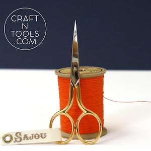 May include: Gold embroidery scissors with a silver blade, resting on a reel of orange thread. The reel has a label that reads "D.C. Granger 410". The scissors are labelled "Sajou" on a white ribbon.