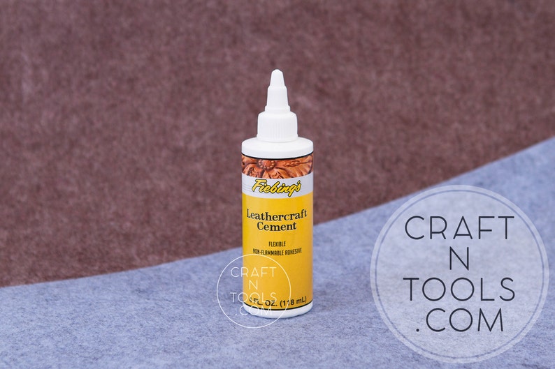 May include: A bottle of Fiebing's Leathercraft Cement, a flexible, non-flammable adhesive. The bottle is white with a yellow label and the text "Fiebing's Leathercraft Cement" is printed on the label. The bottle is sitting on a blue surface.