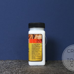 May include: A white bottle of Fiebing's Leather Dye in a clear plastic bottle with a black lid. The bottle is labelled with the product name and instructions for use. The bottle is sitting on a textured surface.