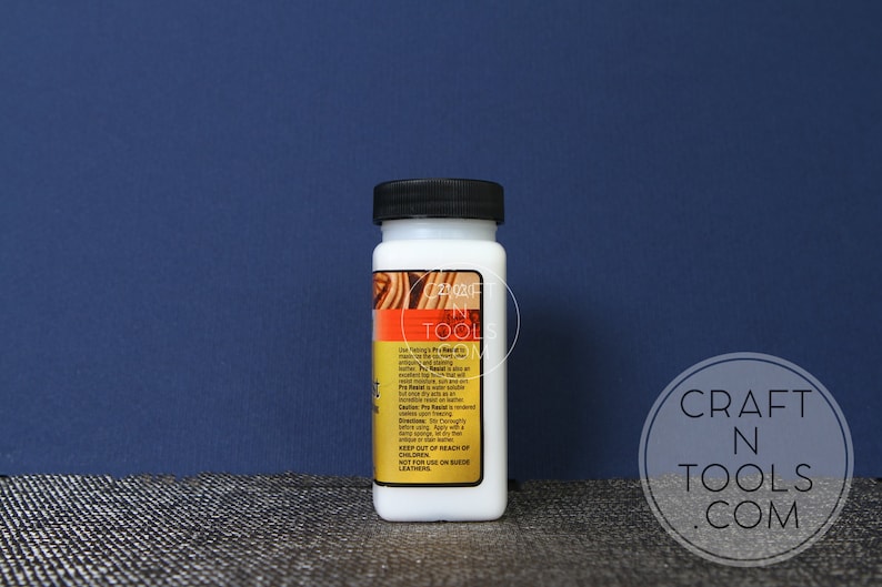 May include: A white bottle of Fiebing's Leather Dye in a clear plastic bottle with a black lid. The bottle is labelled with the product name and instructions for use. The bottle is sitting on a textured surface.