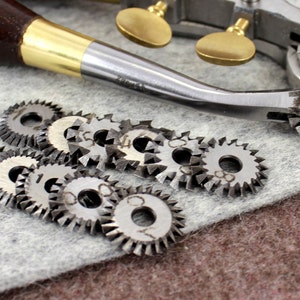 May include: A set of metal rotary blades for a leather crafting tool. The blades are various sizes and have a serrated edge. The blades are on a gray surface.