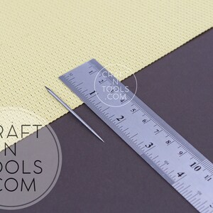 May include: A silver needle for needle felting, measuring 2 inches in length, is placed on a yellow fabric next to a ruler. The ruler is marked in inches and centimeters. The fabric is textured and has a slight sheen. The background is a dark gray surface.