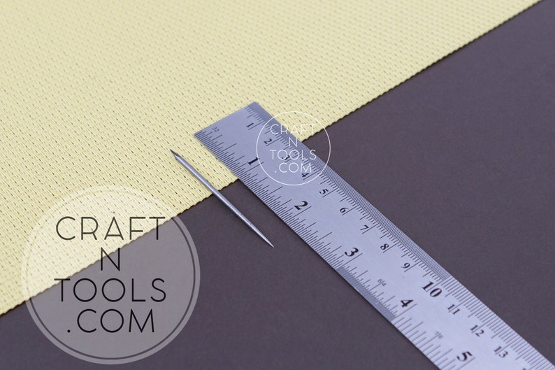 May include: A silver needle for needle felting, measuring 2 inches in length, is placed on a yellow fabric next to a ruler. The ruler is marked in inches and centimeters. The fabric is textured and has a slight sheen. The background is a dark gray surface.