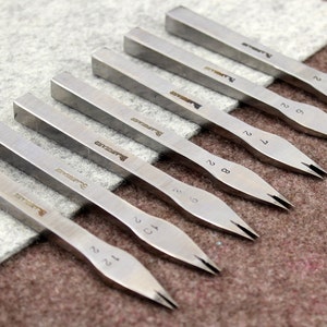 May include: A set of eight silver metal tools with pointed tips and numbered handles. The tools are arranged in a row on a gray fabric surface.