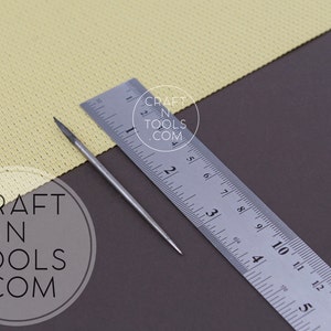 May include: A close-up of a silver metal awl with a pointed tip, measuring approximately 3 inches in length, lying on a brown surface next to a ruler. The ruler is marked in inches and has the text "CRAFT N TOOLS .COM" printed on it.