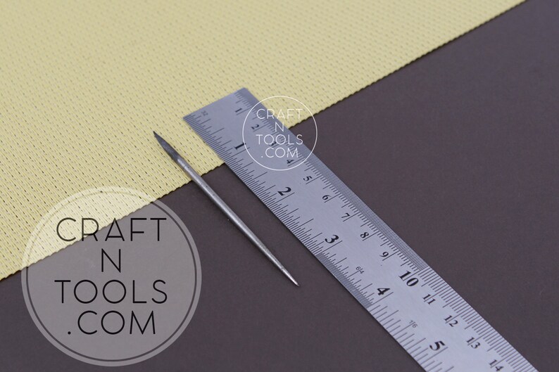 May include: A close-up of a silver metal awl with a pointed tip, measuring approximately 3 inches in length, lying on a brown surface next to a ruler. The ruler is marked in inches and has the text "CRAFT N TOOLS .COM" printed on it.