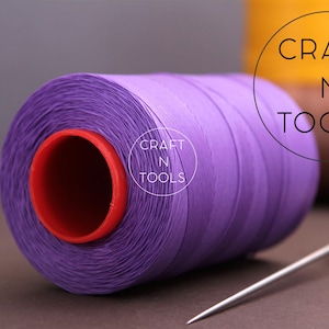 Puede incluir: Una bobina de hilo morado con un centro rojo, etiquetada "CRAFT N TOOLS", junto a una aguja de coser con mango de madera.