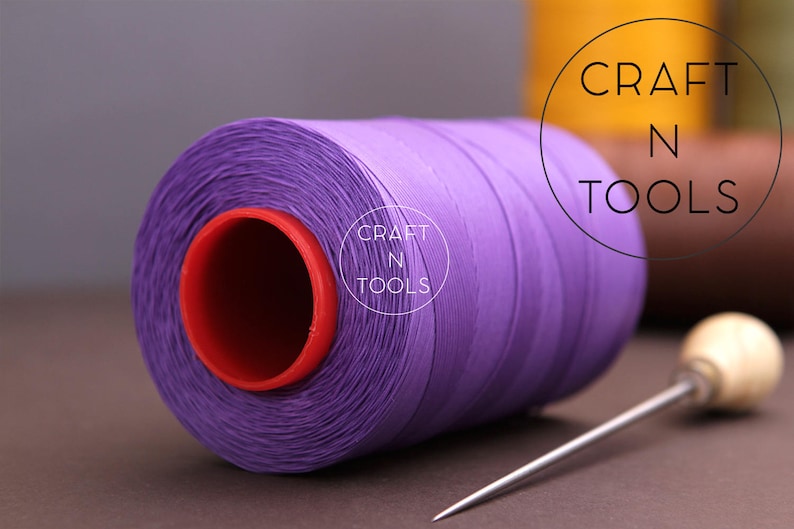 Puede incluir: Una bobina de hilo morado con un centro rojo, etiquetada "CRAFT N TOOLS", junto a una aguja de coser con mango de madera.