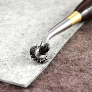 May include: A close-up of a leather crafting tool with a wooden handle and a metal wheel with sharp edges. The tool is resting on a piece of gray felt.
