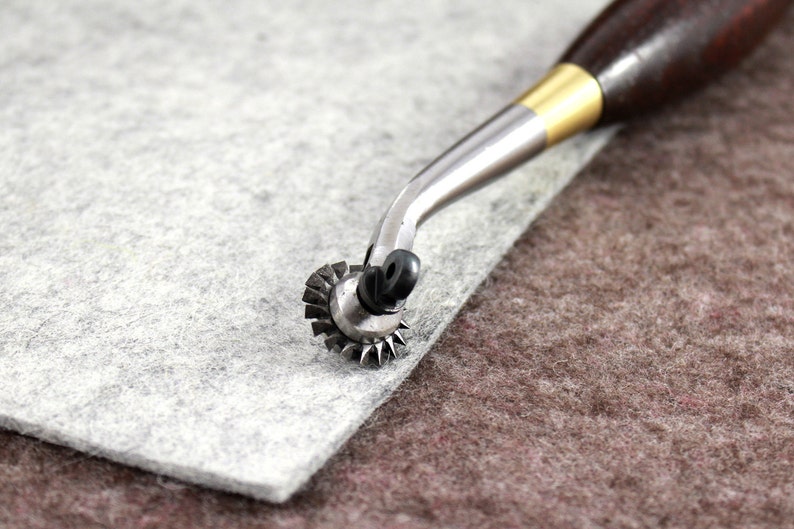 May include: A close-up of a leather crafting tool with a wooden handle and a metal wheel with sharp edges. The tool is resting on a piece of gray felt.