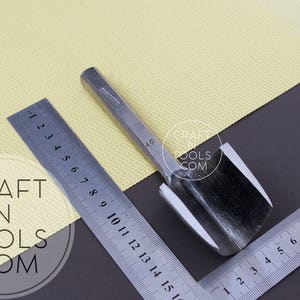 Puede incluir: Una herramienta de metal para trabajar el cuero con un mango y una cuchilla curva. La herramienta est&aacute; sobre una superficie amarilla con una regla al lado. La regla est&aacute; marcada con los n&uacute;meros del 1 al 15.