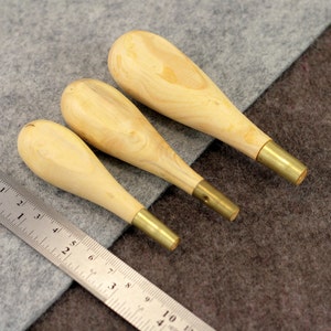 May include: Three wooden awls with brass tips, arranged in order of size, on a gray and brown fabric background. A metal ruler is visible in the lower left corner.