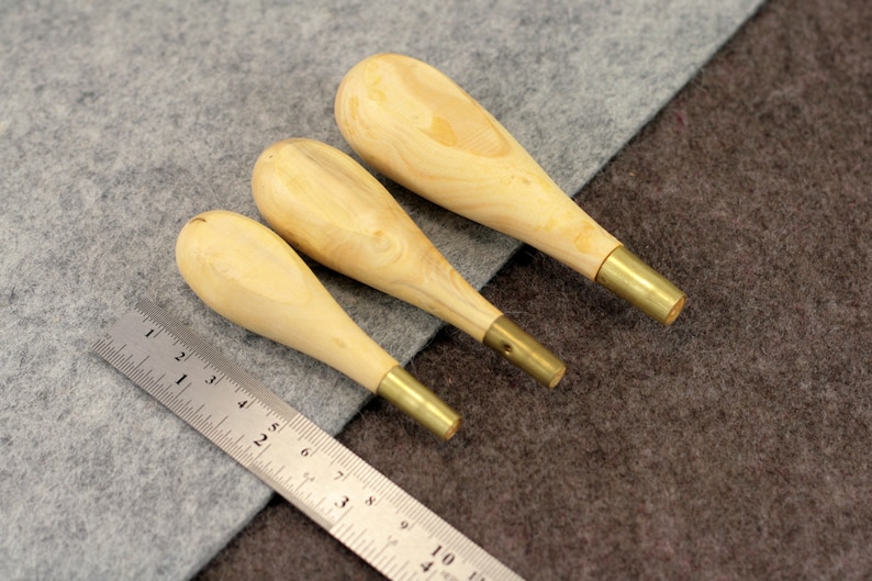 May include: Three wooden awls with brass tips, arranged in order of size, on a gray and brown fabric background. A metal ruler is visible in the lower left corner.